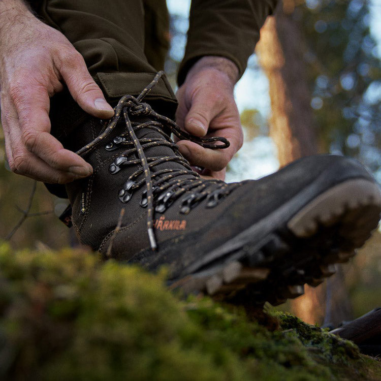 A close-up lifestyle shot of a person tying the laces of a Harkila hiking boot while wearing olive green pants, set against a blurred forest background.