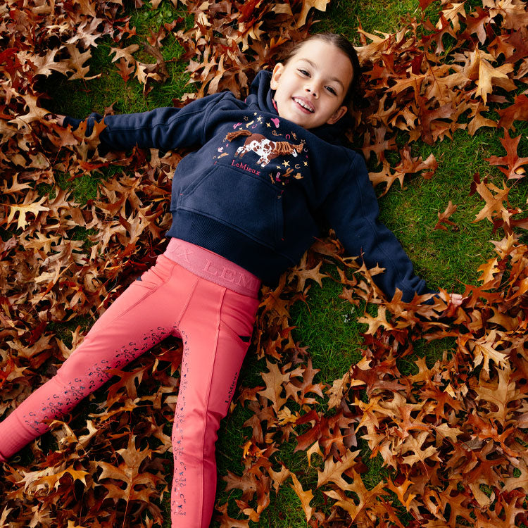 Child lying on a bed of autumn leaves wearing a navy hoodie and pink pants.