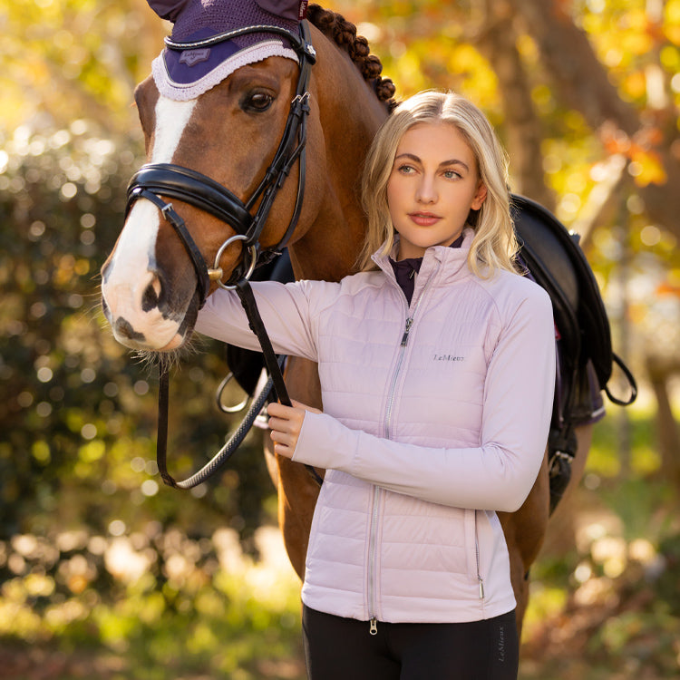 Woman in a light pink LeMieux jacket standing next to a horse with a blurred natural background
