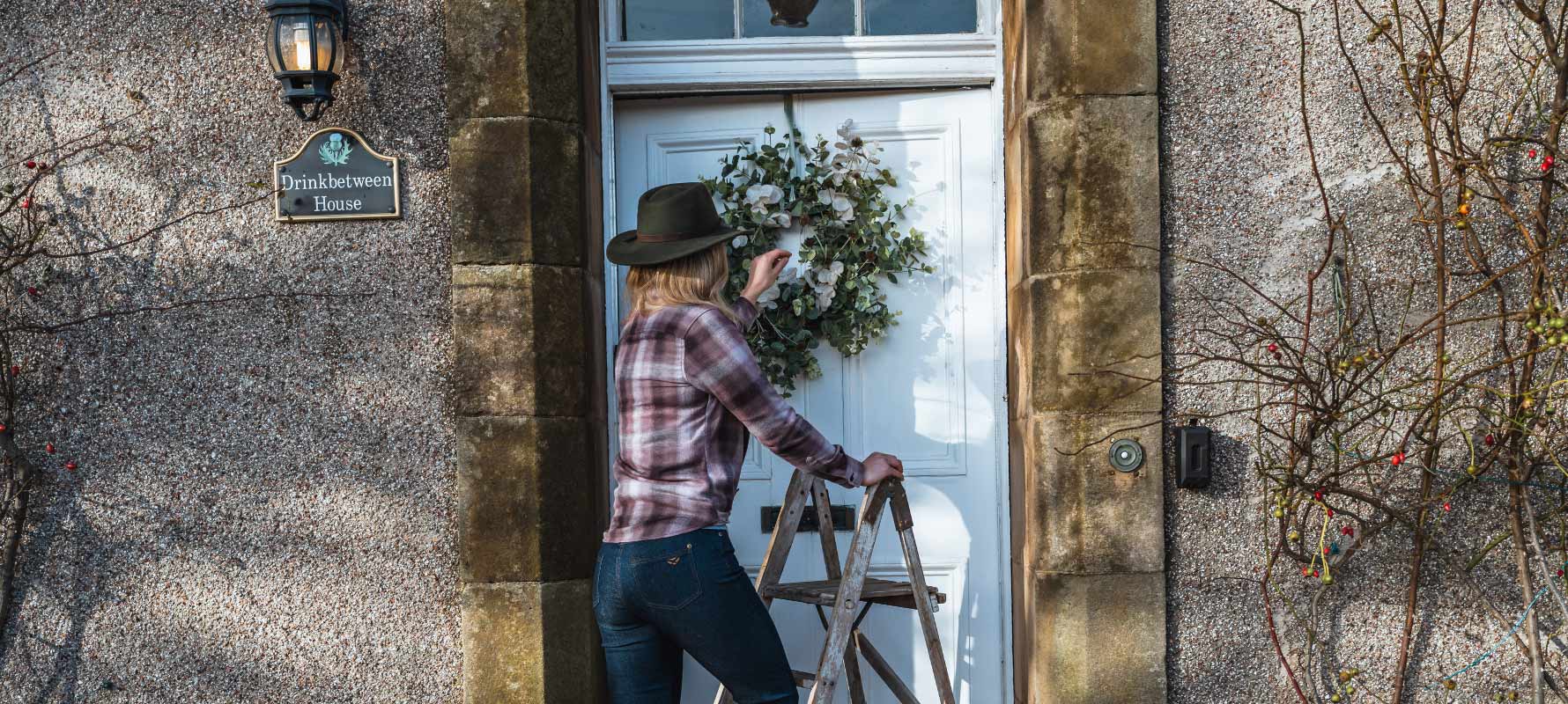 Person with a hat and plaid shirt standing next to a door with a wreath, outside a stone building.