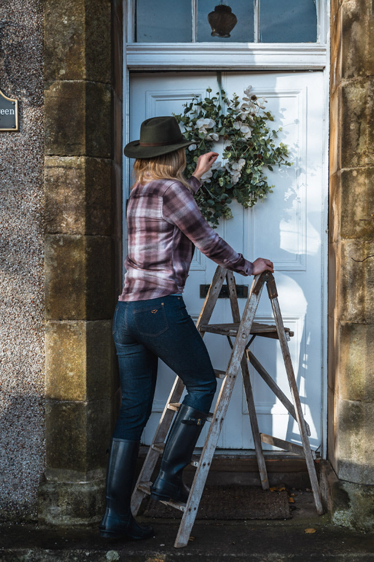 Person with a hat and plaid shirt standing next to a door with a wreath, outside a stone building.