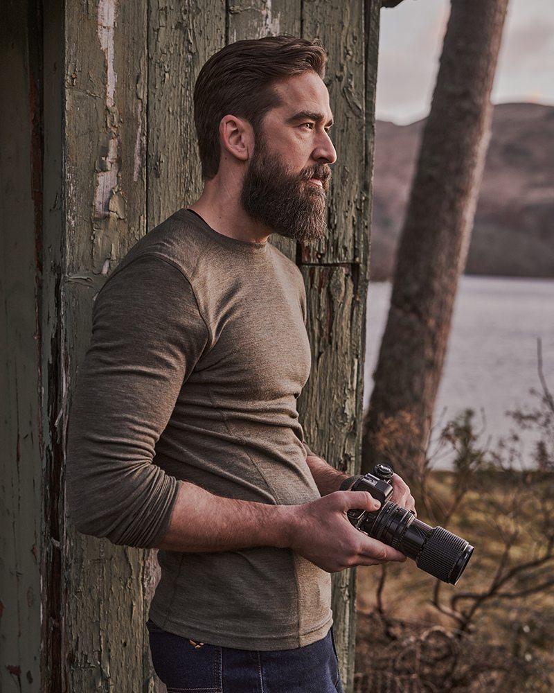 Male model wearing a Hoggs of Fife olive long sleeve base layer outdoors by a lakeside cabin