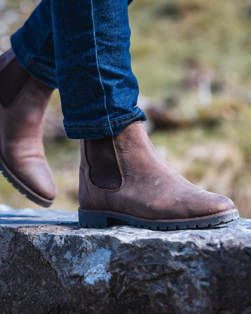 Close up image of Hoggs of Fife brown leather dealer boots being worn to walk on a stone surface with green background. 