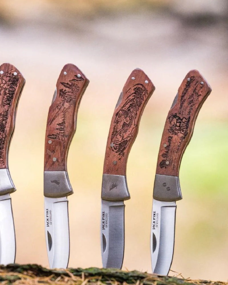 Set of Jack Pyke folding knives with engraved wooden handles displayed outdoors