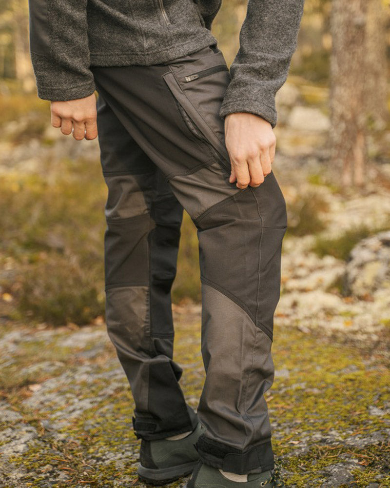 A person outdoors wearing dark gray technical hiking pants with zippered pockets and reinforced knee panels, paired with a heather gray fleece top and boots, set against a blurred forest background.