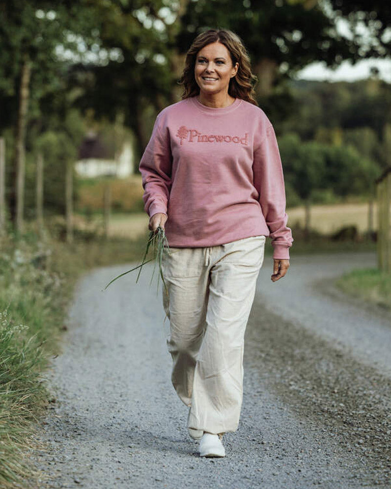 A women walking down a gravel path wearing a pink pullover with light khaki trousers with trees in the background