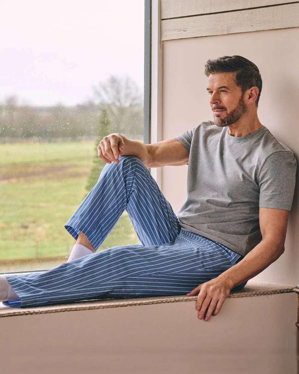 Man relaxing indoors wearing blue striped HJ Hall pyjama bottoms beside a window