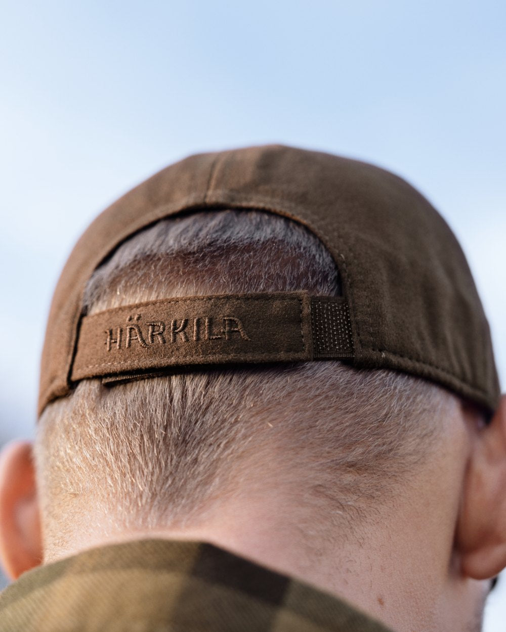 Close-up back view of a person wearing a brown Härkila baseball cap outdoors under a pale blue sky. 