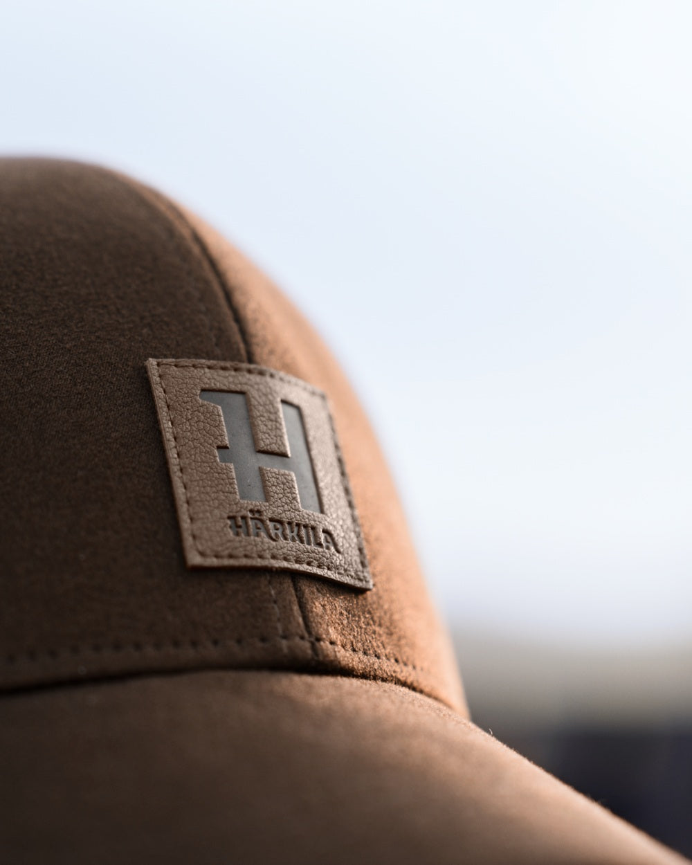Close-up of a brown cap with a rectangular leather patch featuring a large embossed letter H and the brand name. 