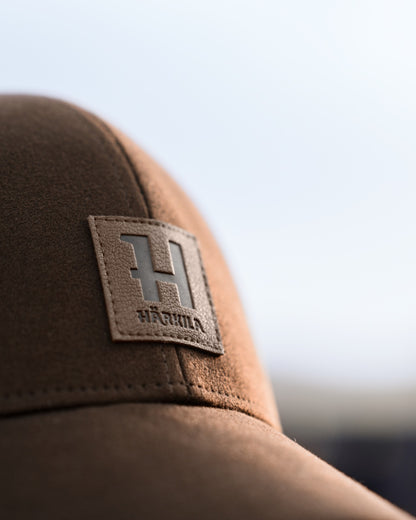 Close-up of a brown cap with a rectangular leather patch featuring a large embossed letter H and the brand name. 