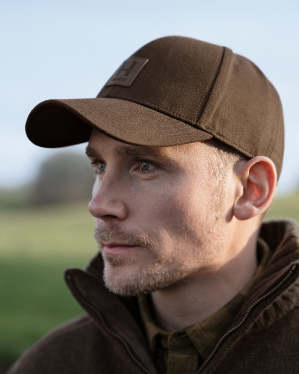 A man in profile wearing a brown baseball cap with a small rectangular logo patch on the front, photographed outdoors against a softly blurred green and blue background. 