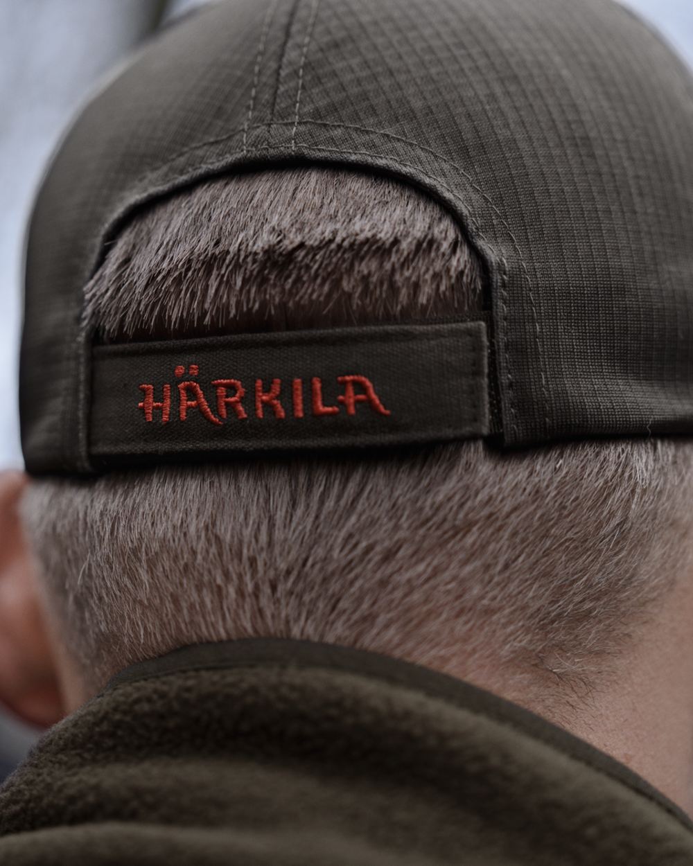 Close-up back view of a person wearing a dark olive green cap, showing the Harkila brand patch in red embroidered lettering on the black adjustment strap. 