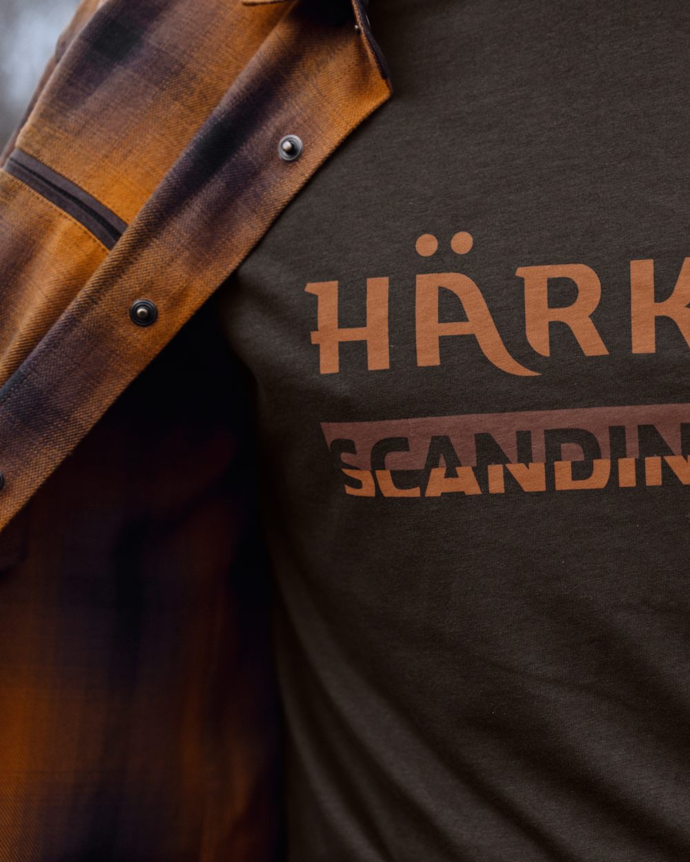 Close-up of a person wearing a dark brown heather t-shirt with a copper-brown graphic print that reads, with an open brown and black plaid flannel shirt draped over the shoulders. 