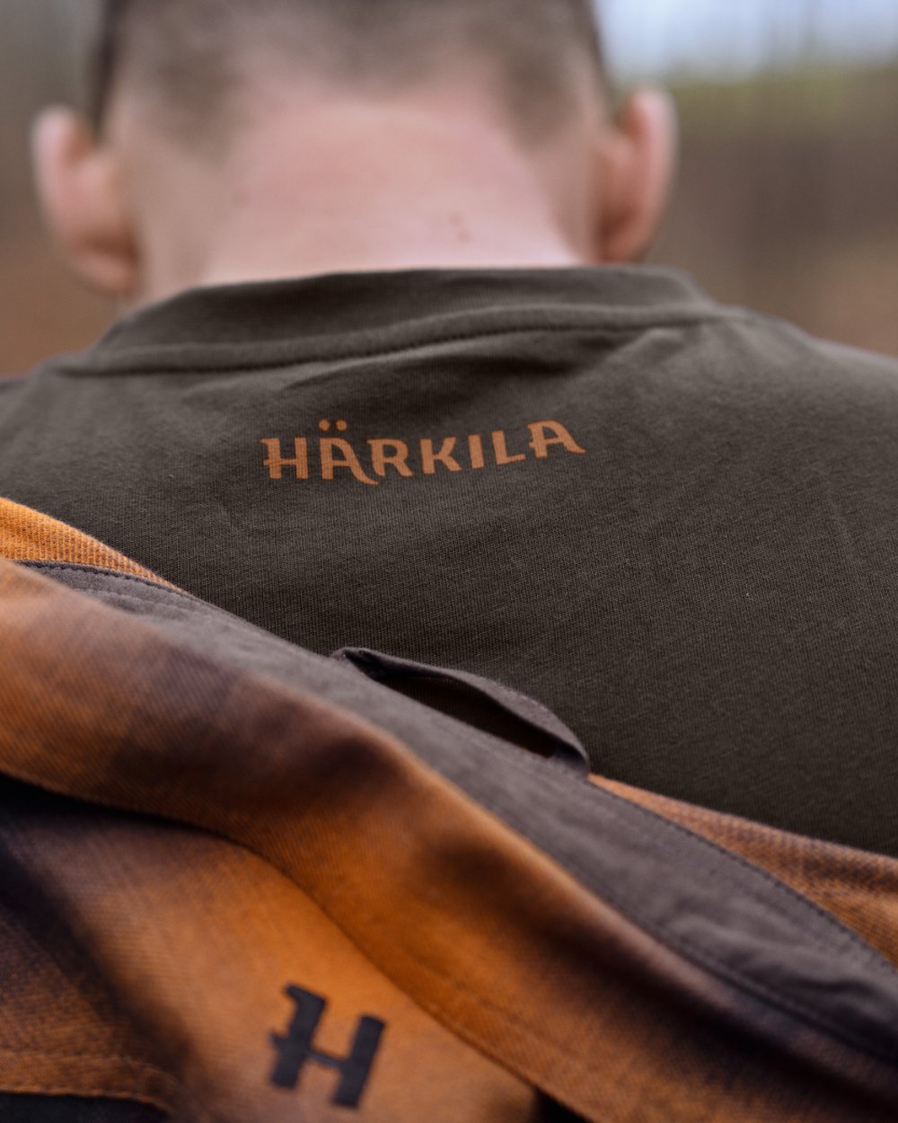 Close-up back view of a person wearing a dark brown Härkila shirt, with a brown and orange plaid scarf draped over the shoulders and the Härkila logo embroidered on the upper back. 
