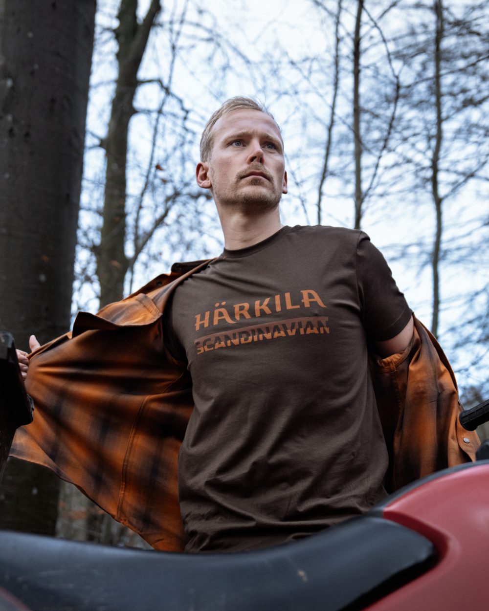 A man wearing a brown t-shirt with printed in orange-brown, holding open an orange-brown plaid flannel shirt in an outdoor forest setting. 
