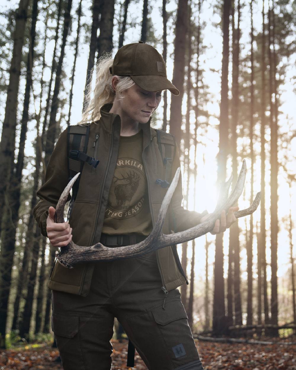 A woman in outdoor hunting apparel stands in a sunlit forest holding a large deer antler. She wears a brown cap, olive green vest, dark olive shirt with a graphic print, and dark cargo pants. 