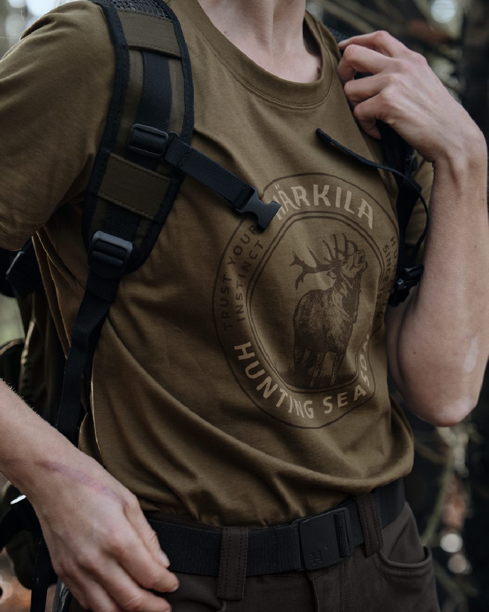 A close-up of a person wearing a brownish-olive tactical t-shirt with a dark brown circular seal graphic featuring an elk and the text &
