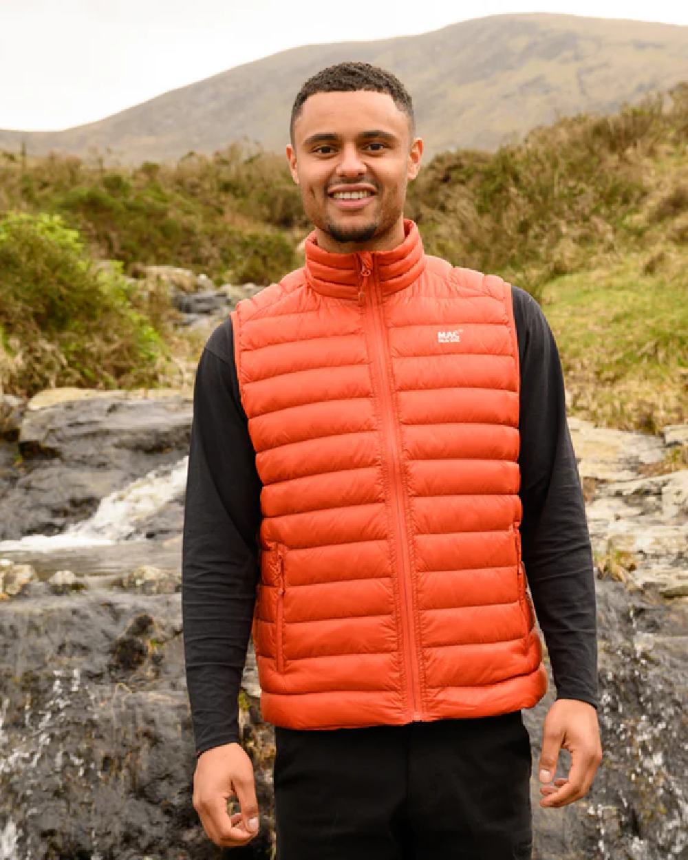 Burnt Orange coloured Mac In A Sac Mens Alpine Down Gilet on blurry mountain background 
