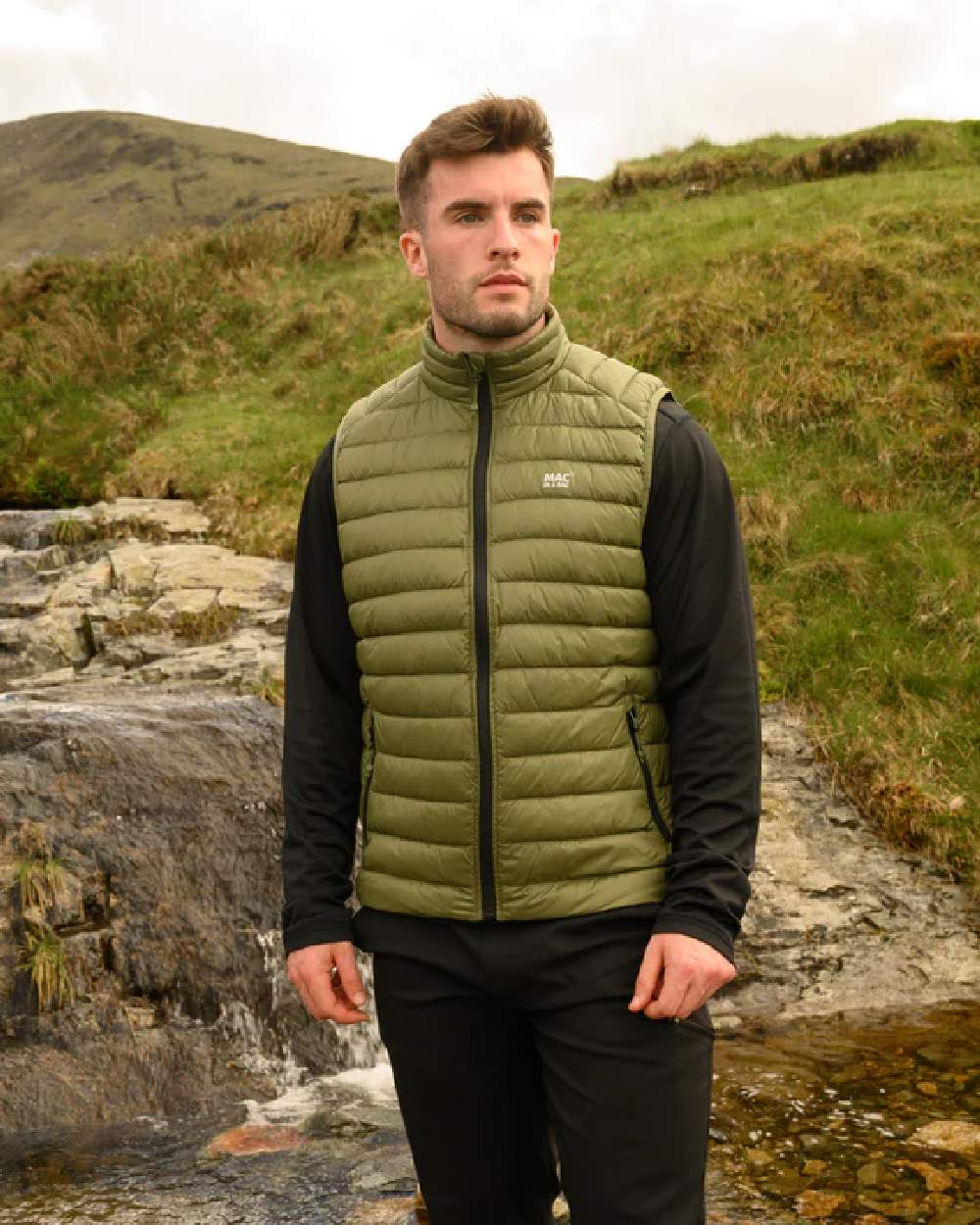 Khaki coloured Mac In A Sac Mens Alpine Down Gilet on blurry mountain background 