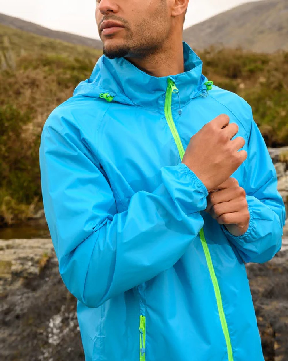 Neon Blue coloured Mac In A Sac Packable Origin Neon Waterproof Jacket on blurry mountain background 