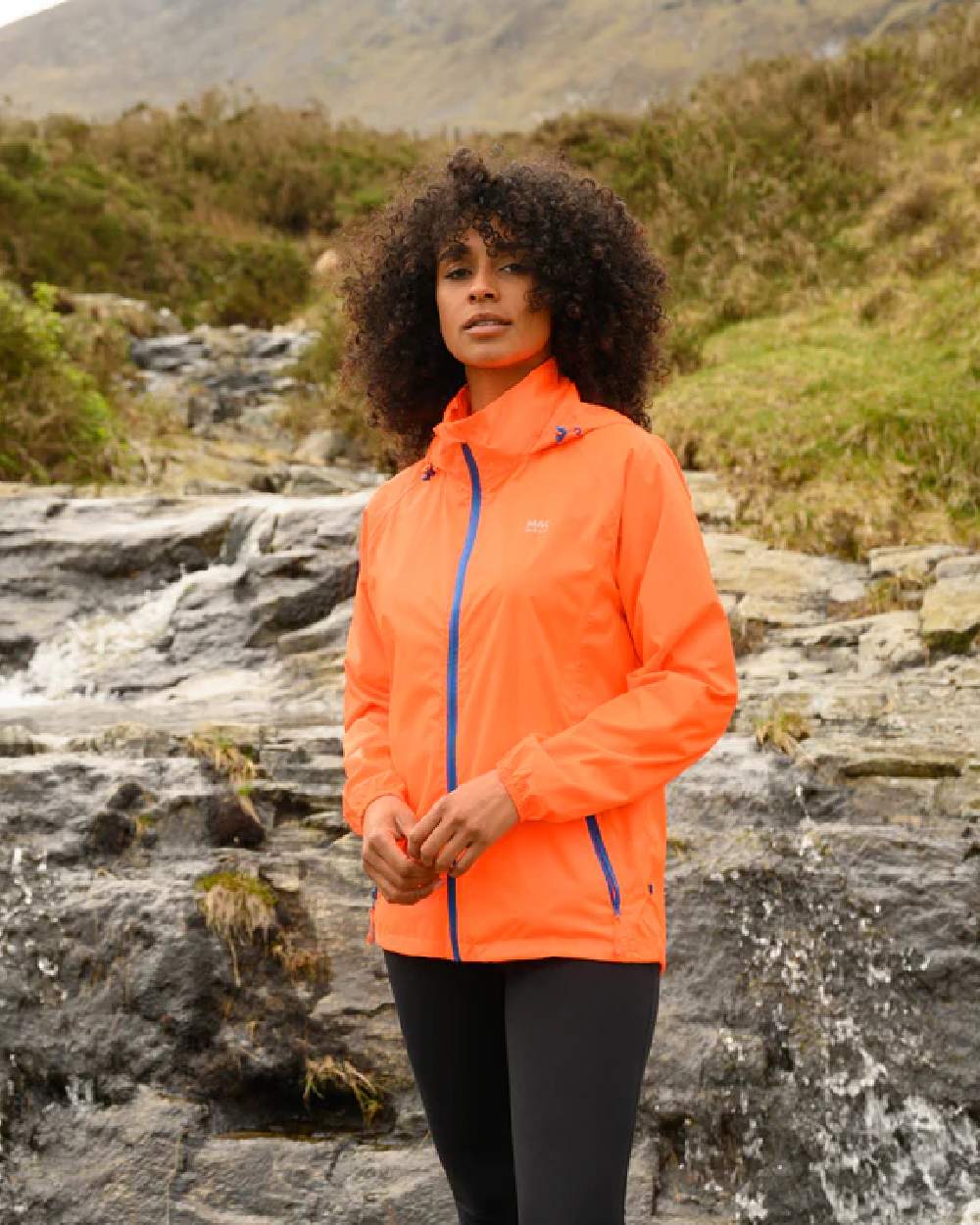 Neon Orange coloured Mac In A Sac Packable Origin Neon Waterproof Jacket on blurry mountain background 