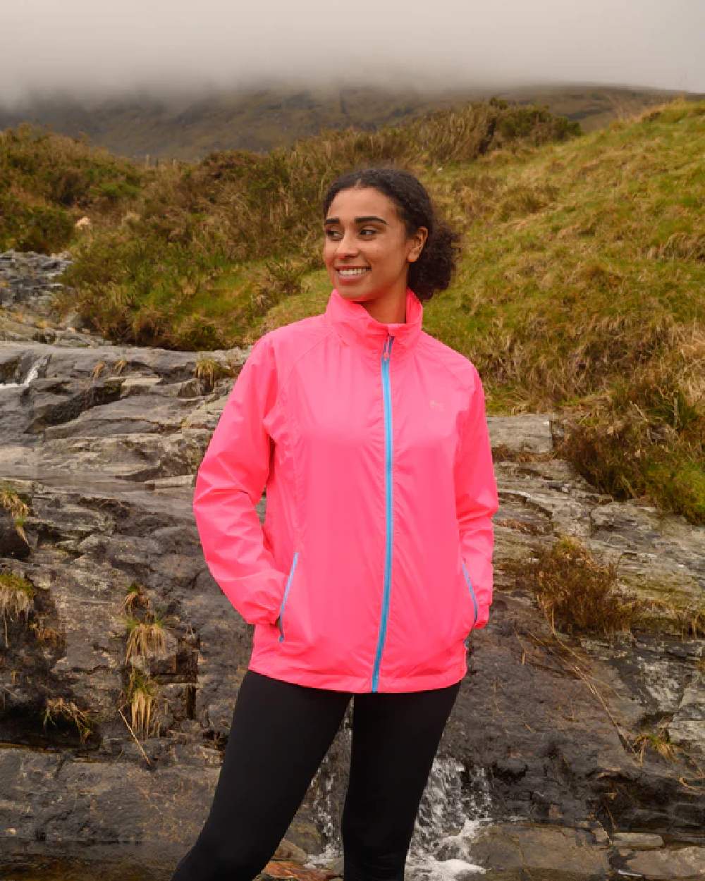 Neon Pink coloured Mac In A Sac Packable Origin Neon Waterproof Jacket on blurry mountain background 