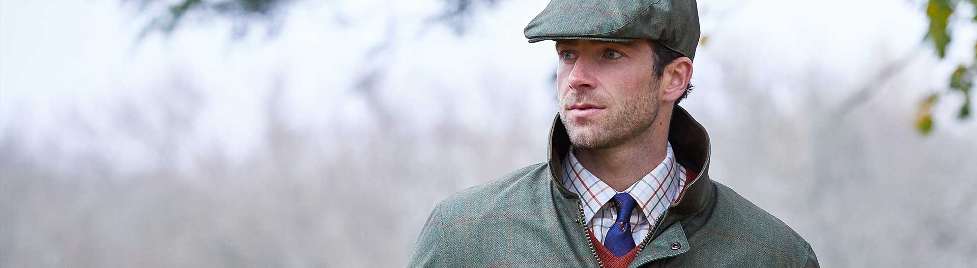 Man wearing Green flat cap and waterproof jacket over a tattersall shirt a blue tie. With blurred grey sky background.