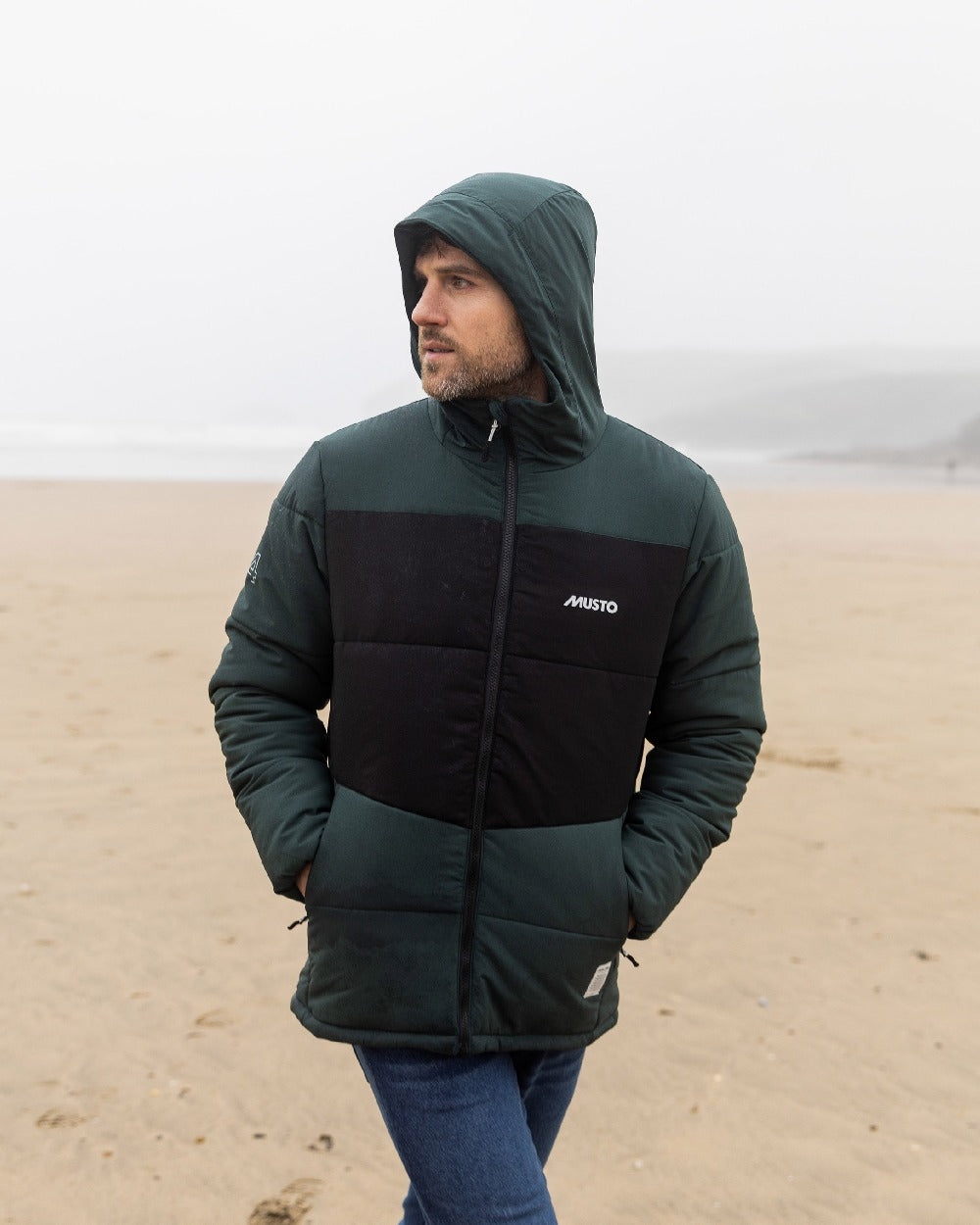 Darkest Spruce Coloured Musto Mens 64 Puffer Jacket on beach background 