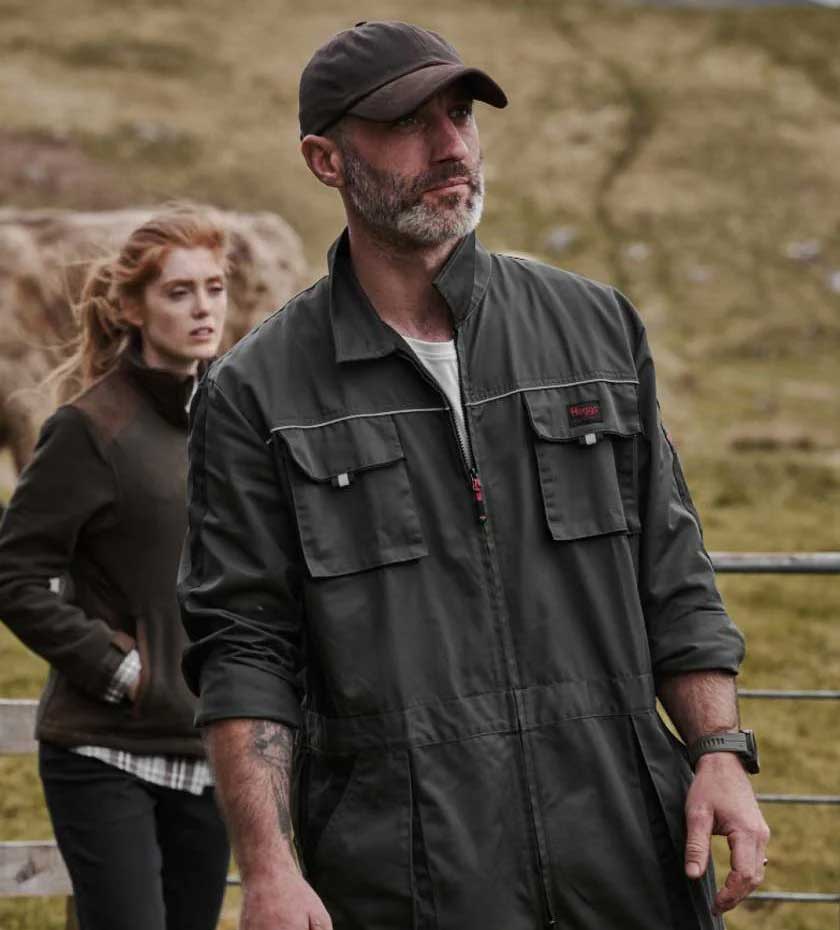 Farmer wearing a green overall with a woman and cow in background.