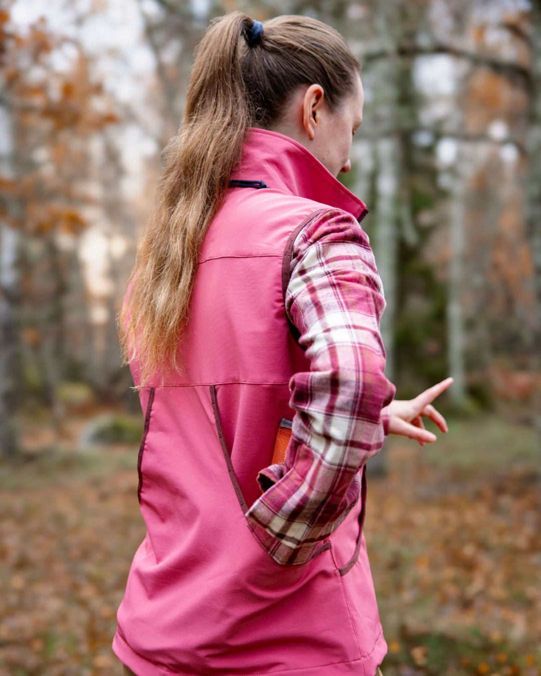 Rear view of a woman with a ponytail wearing a pink Pinewood softshell vest over a pink plaid shirt, standing in an autumn forest with fallen leaves on the ground