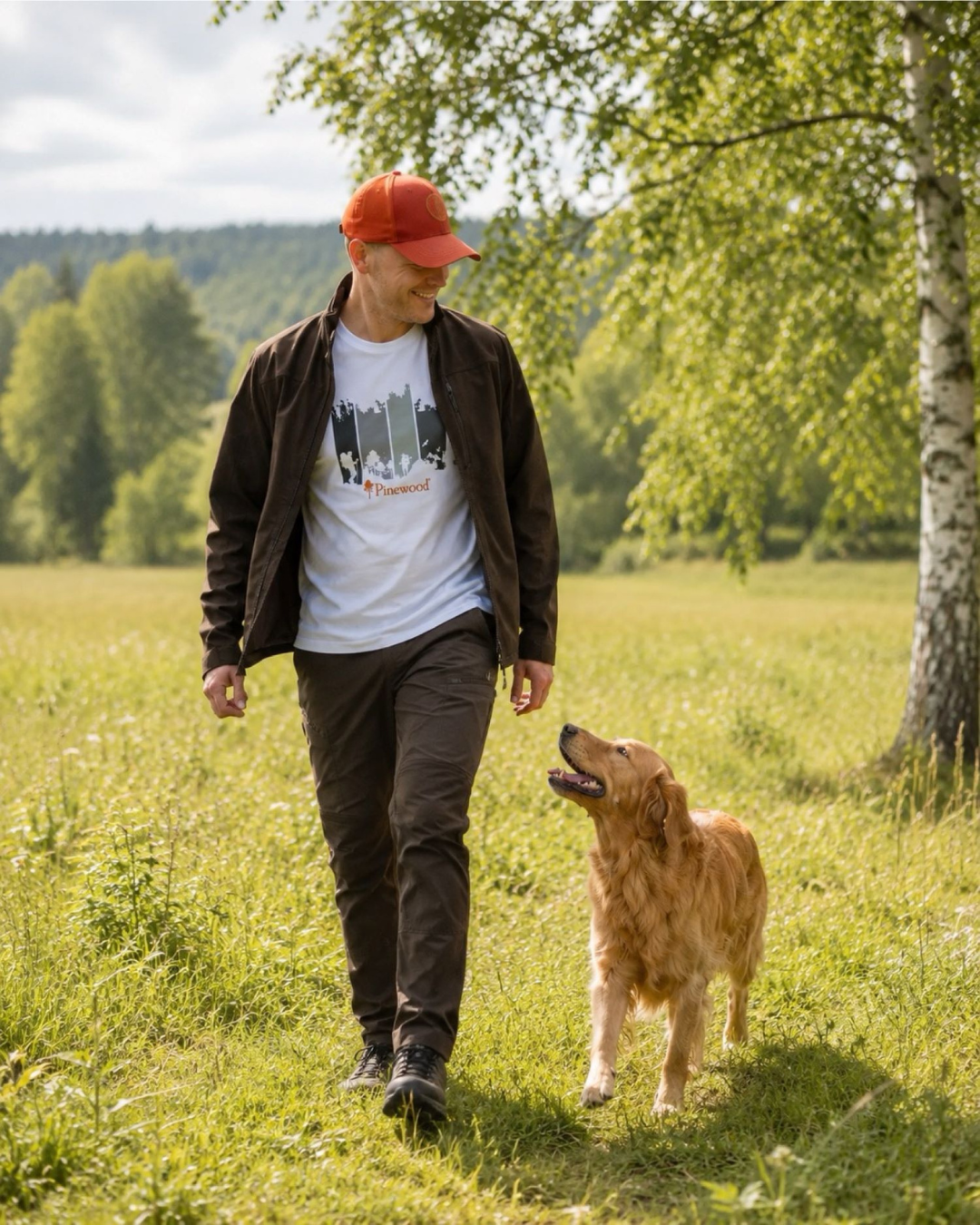Man wearing a red cap, dark jacket, and white Pinewood graphic T-shirt walks through a sunny grassy meadow with a golden retriever, surrounded by trees.