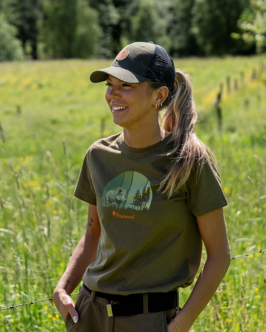 Smiling woman wearing a dark trucker cap and olive green Pinewood T-shirt with a wildlife graphic, standing in a sunlit meadow with a wire fence in the background.