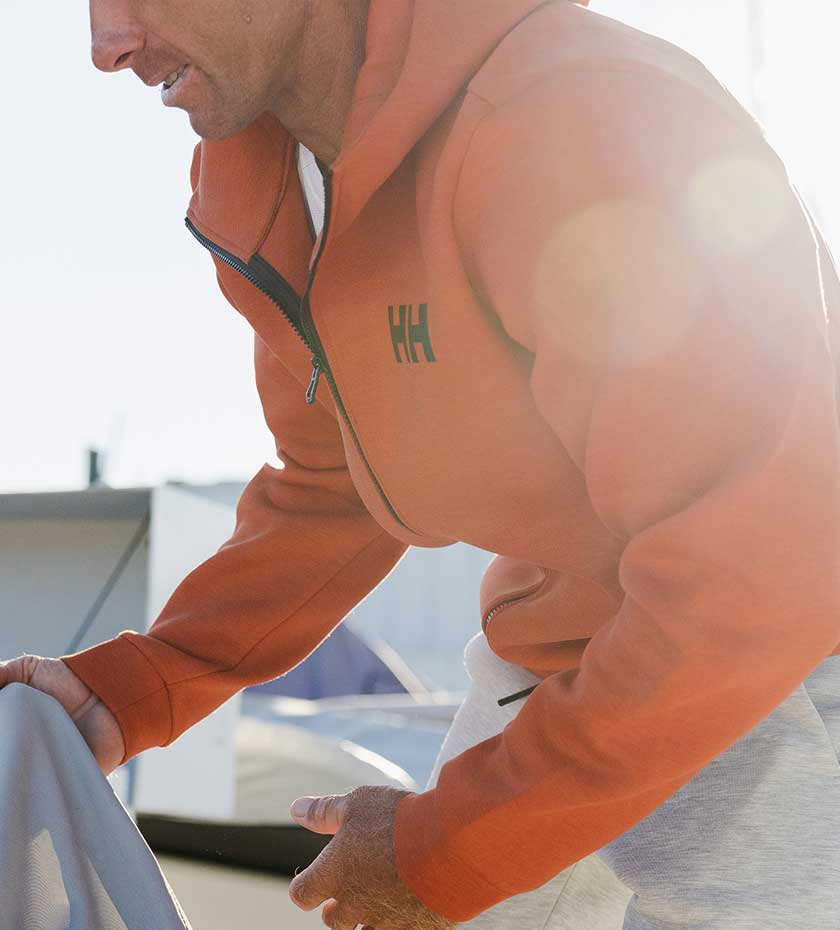 Man in orange HH logoed sailing hoodie lifts a sail on a confusing white background.