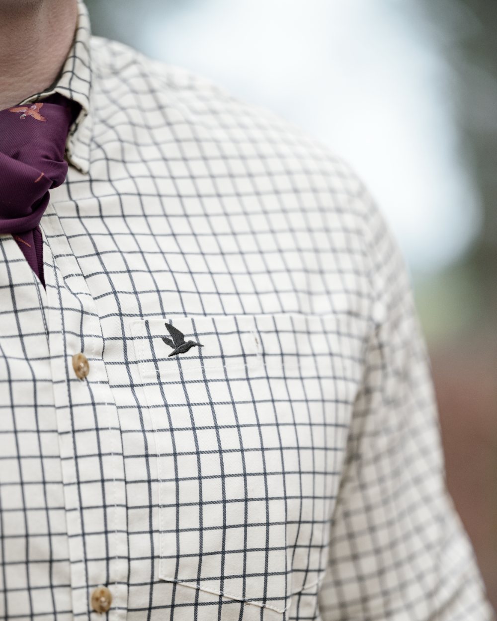 Close-up of a person wearing a white button-down shirt with a dark blue grid pattern, a dark purple necktie, and a small embroidered bird logo on the chest pocket. 