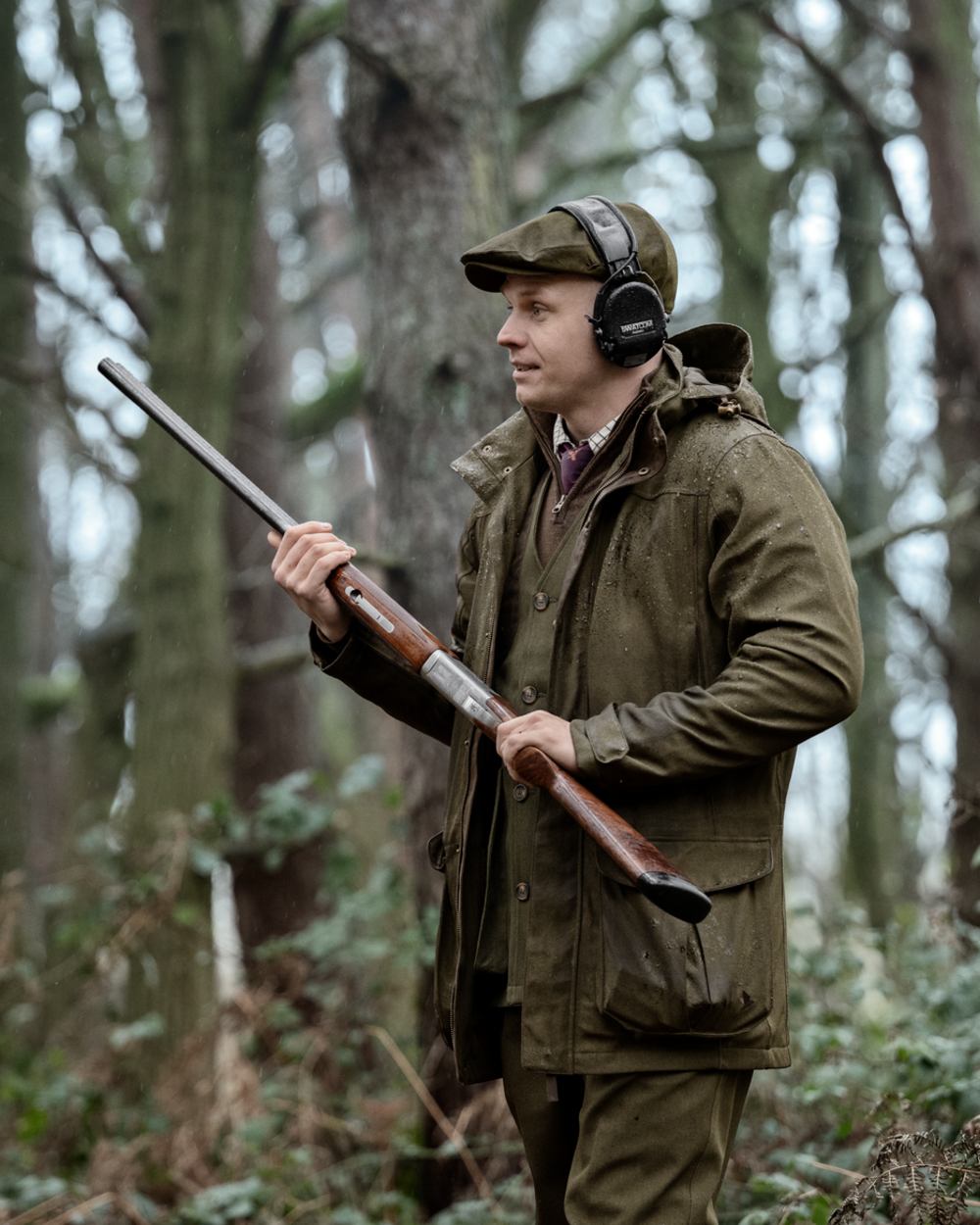 A man in a forest wearing a brown deerstalker hat, over-ear headphones, and an olive green hunting jacket, holding a shotgun. 