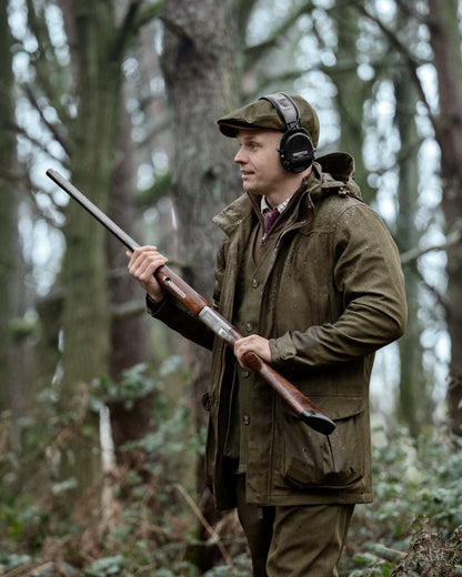 A man in a forest wearing a brown deerstalker hat, over-ear headphones, and an olive green hunting jacket, holding a shotgun. 