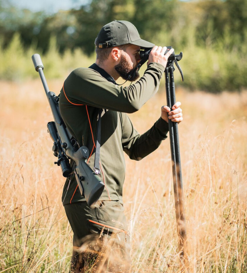 Man in stalking clothing using binoculars in tall grass with rifle on his back