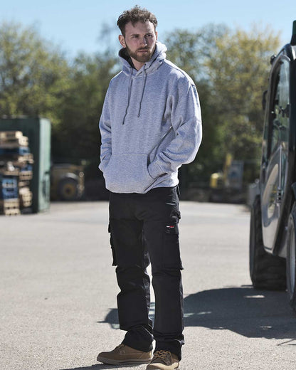 Man in hoodie stands near tractor wearing TuffStuff Pro Work Trousers in Black 
