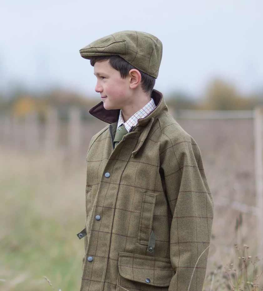 Young boy wears tweed jacket and tweed flat cap with blurred countryside background.