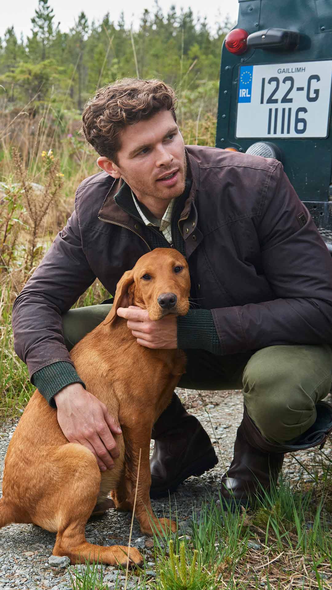 Man in dark brown waxed jacket petting a brown dog behind a Land Rover Defender.