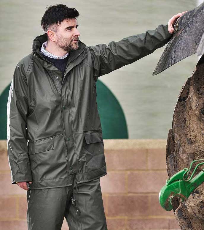 Farmer's waterproof clothing. Man wears Green waterproof jacket and trousers, beside tractor wheel.