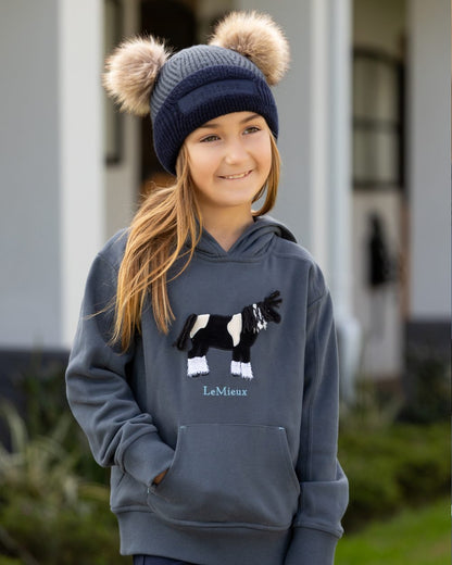 Petrol Coloured LeMieux Mini Clara Beanie On A Outer Yard Background 