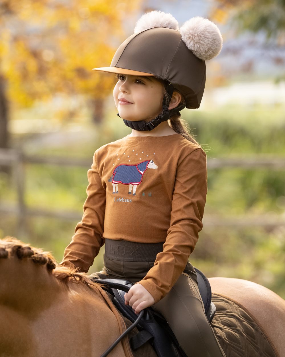 Alpine Coloured LeMieux Mini Double Pom Hat Silk On A Outer Yard Background 