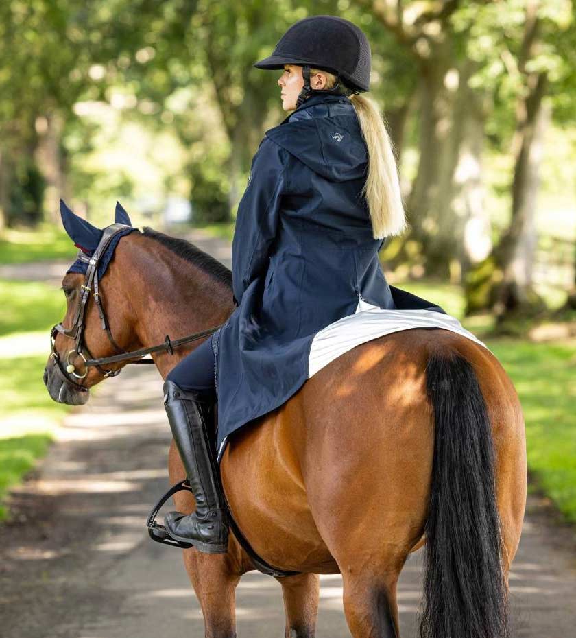 Lemieux jackets - a long navy blue riding coat is worn by a woman on a brown horse.