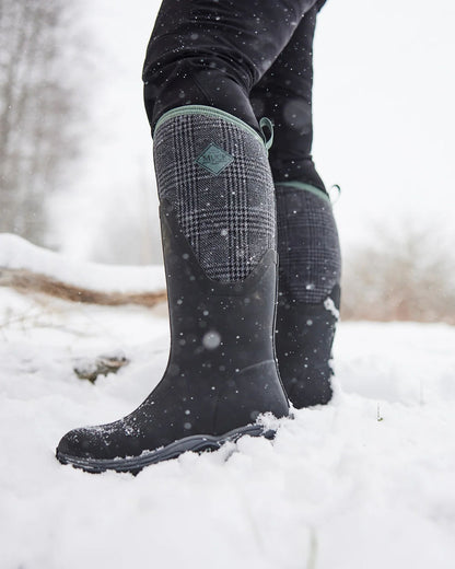 Black Plaid Print Muck Boots Womens Artic Sport II Tall Wellingtons on Snowy background 