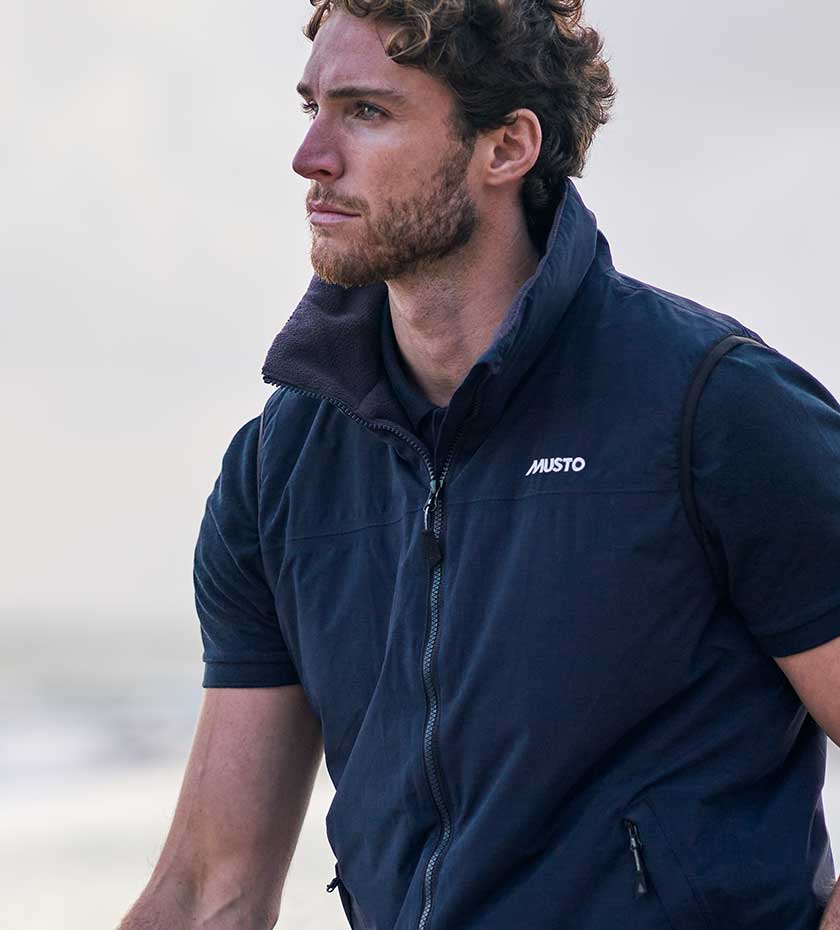 Man wears Musto snug gilet in navy with grey sky background.